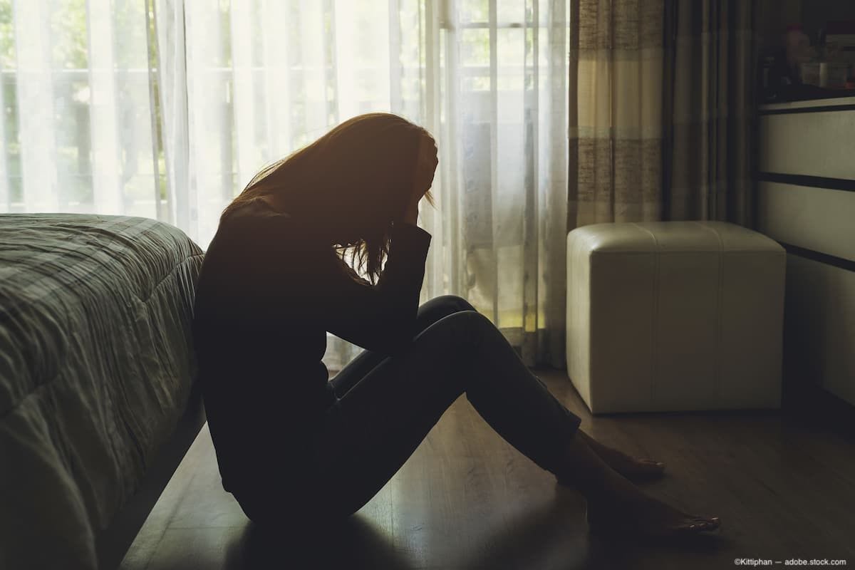 Depressed woman sitting on the floor at the foot of a bed with her head in her hands Image Credit: AdobeStock/Kittiphan