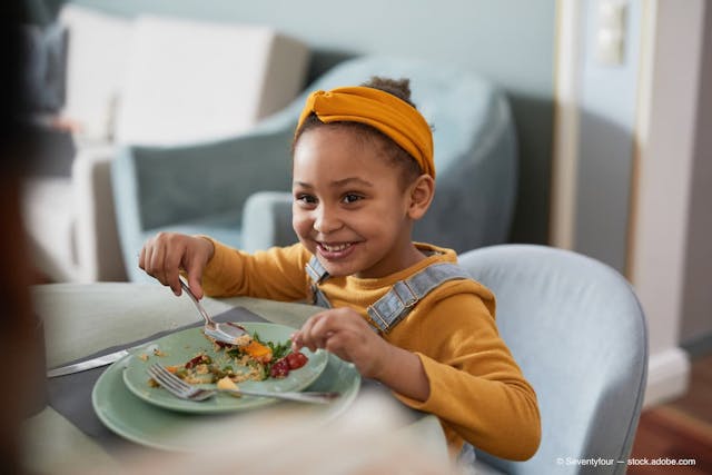 Portrait of cute African-American girl eating food at table and smiling happily while enjoying dinner with family (Adobe Stock / Seventyfour)