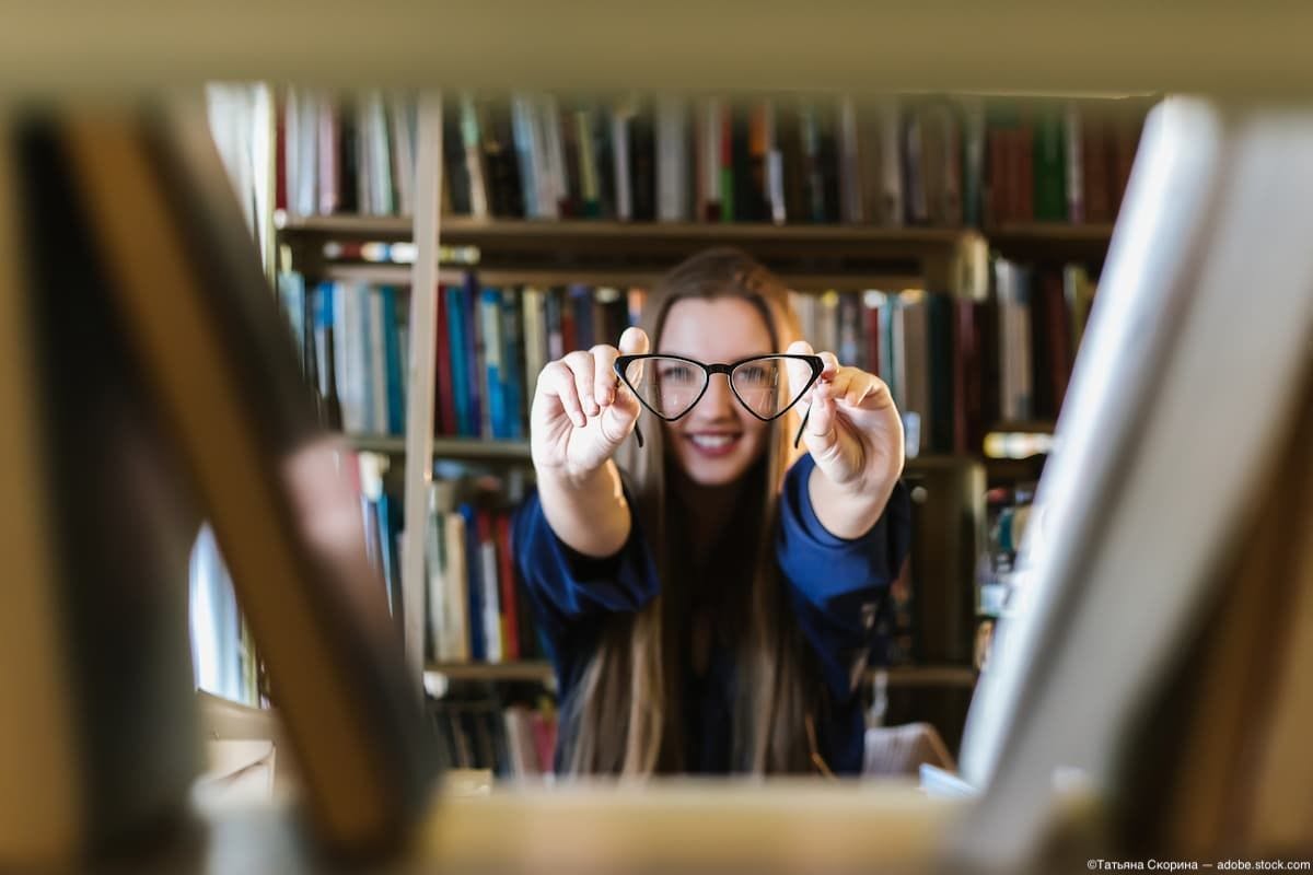 Woman holding up glasses between book shelves in library Image Credit: AdobeStock/ТатьянаСкорина