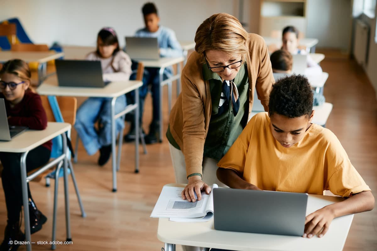 African American schoolboy uses laptop with help of his teacher during computer class in classroom. (Adobe Stock / Drazen)
